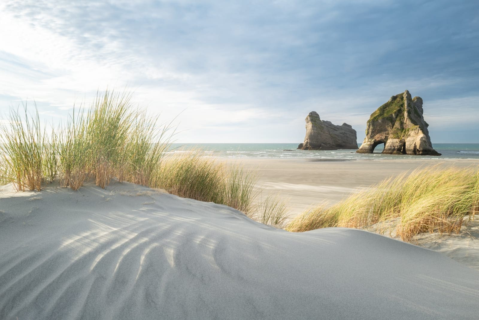 Wind-rippled sand dunes with marram grass at Wharariki Beach, the Archway Islands sea stacks standing offshore under a bright sky.